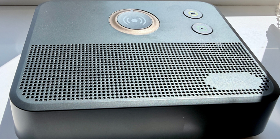 Close-up of a black telecare base unit sitting on a windowsill. The device features a large central emergency call button with a wireless signal icon, two smaller indicator buttons, and a perforated speaker grille covering most of the front surface.