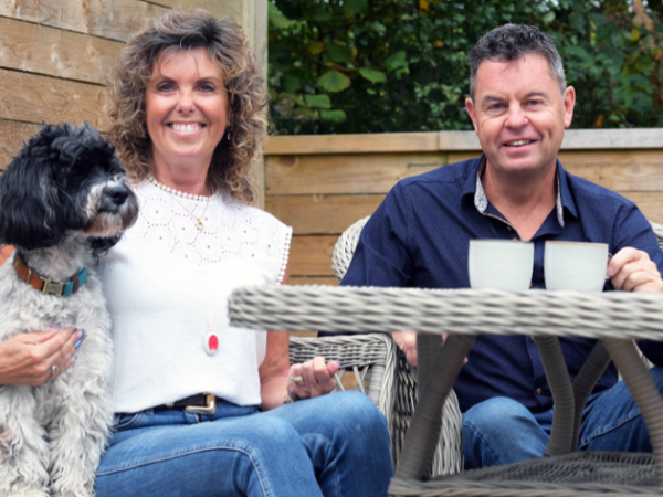 Two people are sitting outdoors on wicker garden chairs beside a table with two mugs. One person is holding a black and white dog sitting on their lap. The setting appears to be a patio or garden area with wooden fencing and greenery in the background.