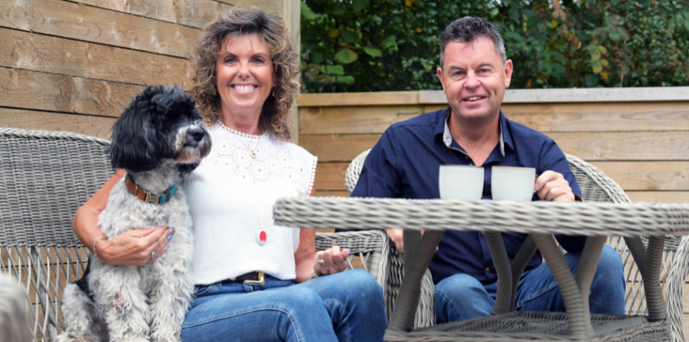 Two people are sitting outdoors on wicker garden chairs beside a table with two mugs. One person is holding a black and white dog sitting on their lap. The setting appears to be a patio or garden area with wooden fencing and greenery in the background.